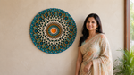 A woman in a light beige and orange floral saree stands next to a large, circular, hand-woven mandala wall hanging in teal, white, and orange. The scene is set against a warm beige textured wall with a hint of green foliage on the right. image 10
