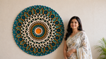A woman in a white floral saree stands beside a large circular, mandala-style wall hanging with teal, cream, and orange geometric patterns against a beige wall. image 13