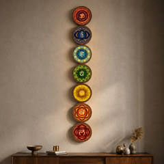 A vertical arrangement of seven circular chakra wall art pieces in red, orange, yellow, green, blue, and brown tones, mounted on a textured beige wall above a wooden console with decorative objects.
