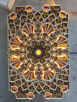 An intricate 3D laser-cut mandala wall art piece with layered geometric patterns, illuminated by warm internal lights. The design features shades of brown, gold, orange, and white, with a bright glowing yellow center, photographed from directly above on a gray concrete floor. image 3